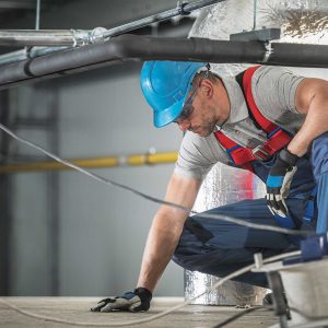 Caucasian HVAC Worker in His 40s Wearing Professional Safety Harness Working Inside Warehouse Building Air Circulation System. Heating, Cooling and Air Filtering.